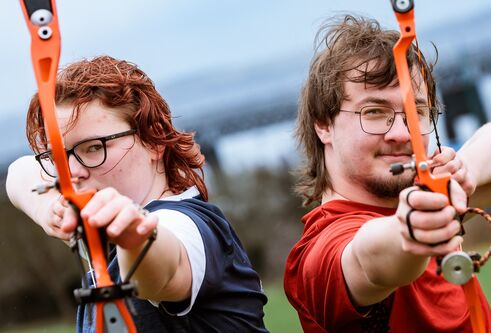two students standing back to back, each holding an archery bow and pointing it forwards
