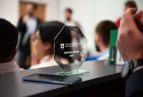 Venture 2026 competition trophy in the foreground with some people blurred out in the room behind it.