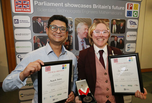 Two people stood smiling holding framed certificates stood in front of a pop up banner. One individual is also holding a medal in a box.