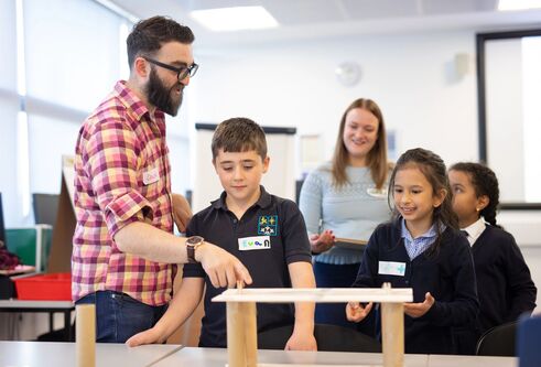 A teacher wearing glasses and a plaid shirt guides a group of schoolchildren as they build a small wooden structure on a table in a bright classroom
