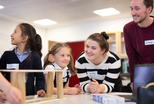 Two adults lean over a table smiling as young children in school uniforms observe and interact with a simple wooden construction during a classroom activity