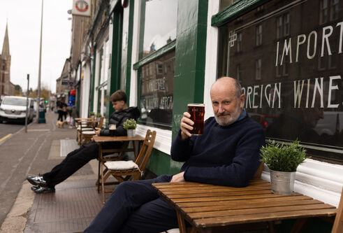 A smiling Fred Macaulay sits at a bistro-style table outside the Speedwell Bar - better-known as Mennies - on Perth Road in Dundee, Scotland. He holds a pint of beer up in a "cheers" gesture
