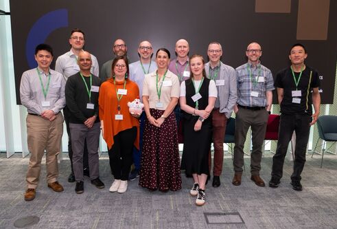 Group photo of researchers and staff wearing name badges, standing together at a conference or showcase event, with presentation screens in background