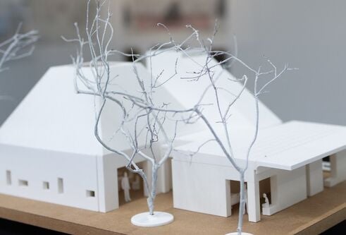 miniature model of a large house made from white material. Tiny little figures stand nearby and there are small branches outside to resemble trees in the winter without their leaves