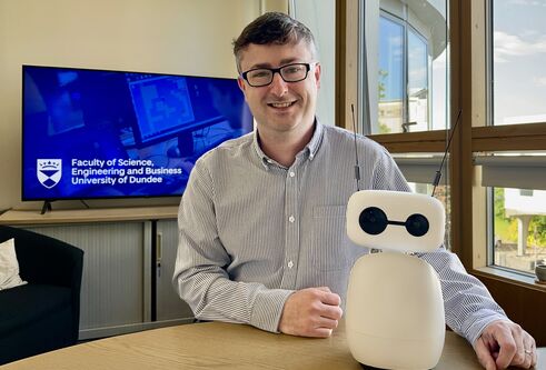 Dr Mike Crabb sitting at a table with a small, white robot, with black camera eyes and two black antenna. In the background is a television screen showing a blue backgrond with University of Dundee branding.
