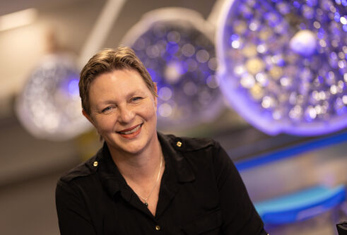Professor Lucina Hackman in black coloured jacket, smiling. Behind her are three large, circular lights, which are tinged with purple.