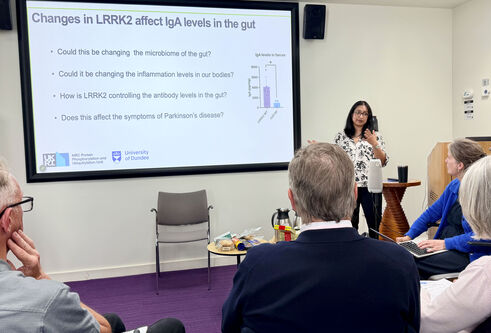 A woman stands at a large smart board, presenting to people sitting down in a room.