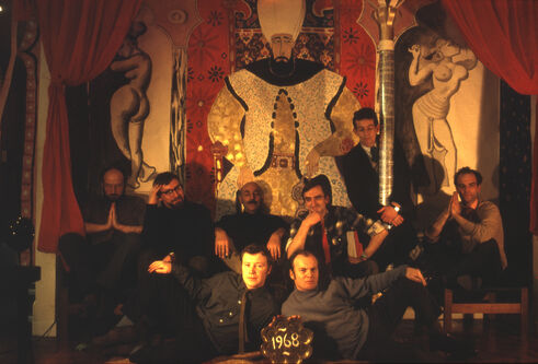 Group portrait of nine people posing before a large ornate mural with theatrical red curtains and a "1968" sign in front.