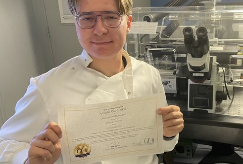 A man wearing a lab coat sat in front of a confocal microscope holding a certificate