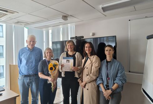Esther Sammler receiving her award and a presentation of flowers from well-wishers, standing in a classroom environment