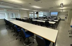 A room full of desks and blue chairs with wheels. There are windows along the left wall.
