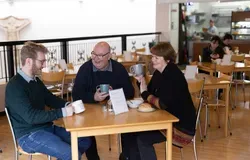 Three people sit around a wooden table smiling and drinking from mugs.