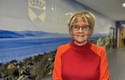 Professor Jill Belch, in a red jumper, standing in front of a blue background with the University of Dundee branding