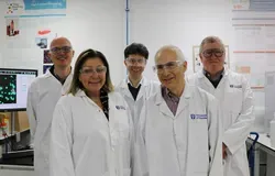 five scientists wearing lab coats and safety glasses, standing next to computer and lab equipment, looking and smiling at the camera
