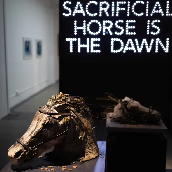 A horses head in bronze and a neon sign at the back with text Sacrificial Horse is the Dawn as part of the Degree Show 2025 exhibition