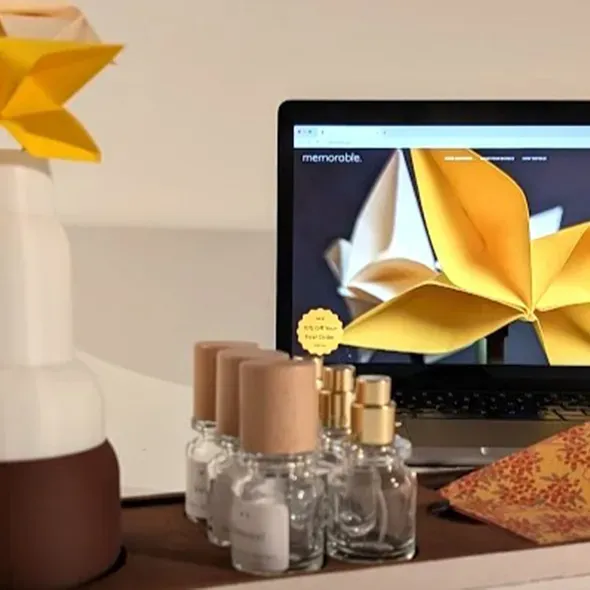 Degree show display with folded paper flower designs, perfume bottles and a laptop showing matching branding on a minimalist table
