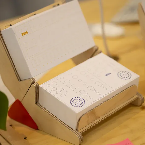 Wooden display stand holding Korg product mockup boxes with minimalist control panel illustrations on a wooden table