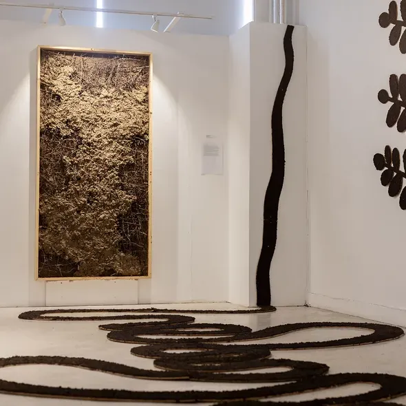 Art gallery installation with earthy abstract artwork, looping dark forms on the floor, and a tree-like wall sculpture.