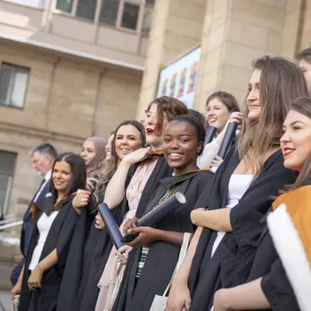 Graduates with certificats and wearing full roves standing on steps and smiling to have their photo taken
