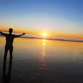 Person standing on beach at sunset with hands raised