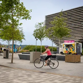 People sat a picnic tables outside V&A Dundee as cyclists go past