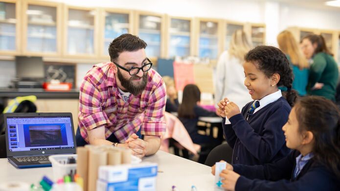male student with two primary school girls, building something out of cardboard