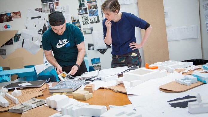 A student consults a book with a lecturer standing beside him, the table contains more books and model buildings