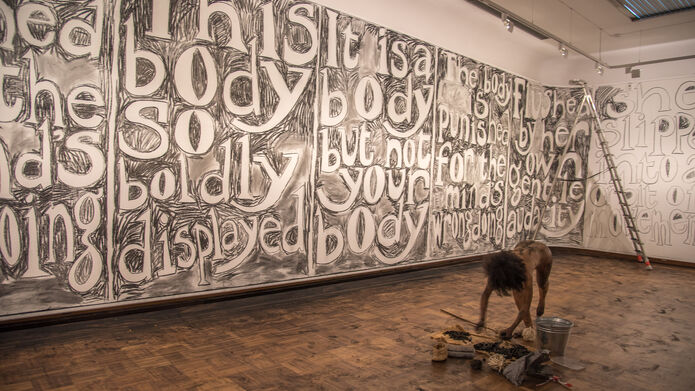 Large words are written in charcoal on a gallery wall. An artist is in the space without clothes working on the drawn lettering. The charcoal soot is everywhere in the space. 
