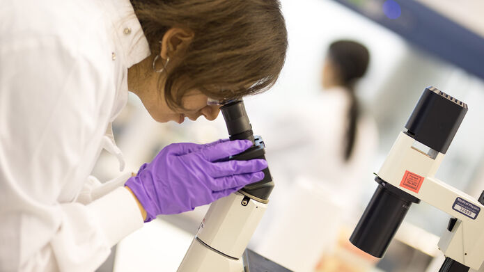 woman in lab coat with purple gloves looking into a microscope