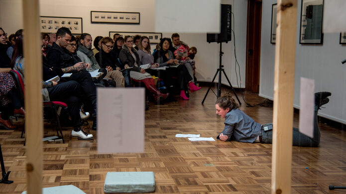 A group of people listening to a person who is lying on a parkay floor reading a book