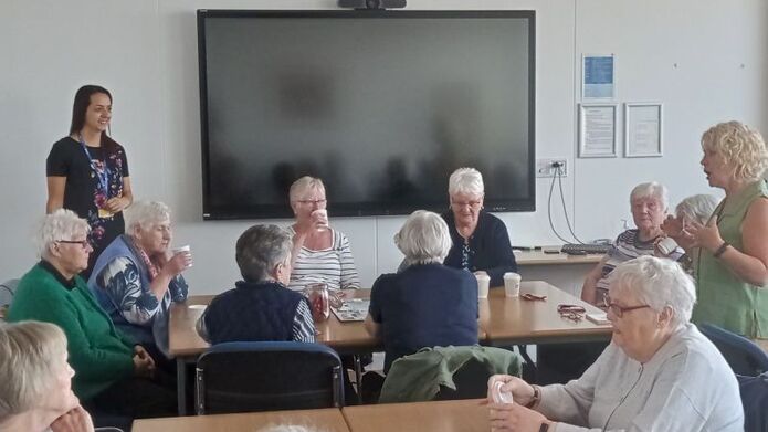 A group of older women from Dundee sit around tables holding takeaway coffee cups as they listen to a woman speaking.