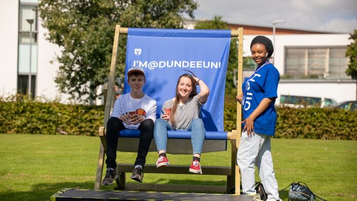 Three young adults sitting together on a University of Dundee branded lawn chair, enjoying leisure time in a grassy area under clear skies.