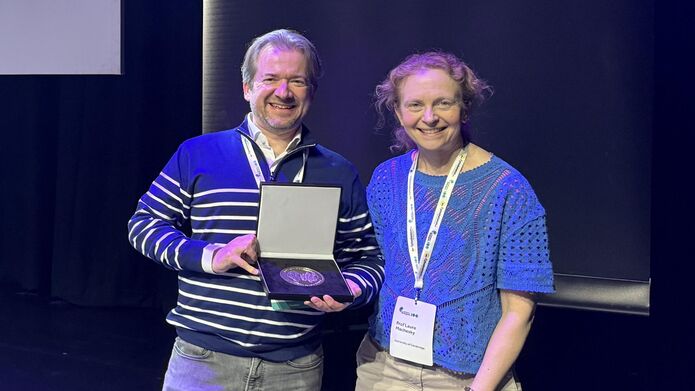 Man and woman standing next to each other both smiling and looking at the camera. Man is holding a large medal in an open box.