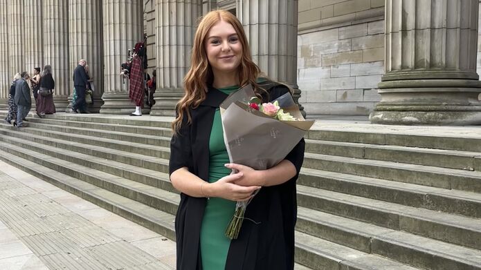 A person stands on the steps outside of a building with large pillars, they are wearing graduation robes and a green dress, holding a bunch of flowers.
