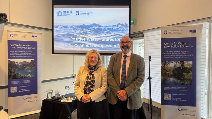 A man and woman stood smiling in front of a projector screen and 2 banners