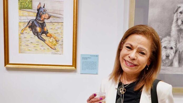 A brunette woman stands in a gallery with a glass in her hand, in front of a painting of a Doberman in a gold frame.