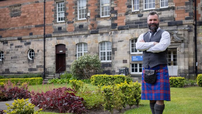 A man in a kilt stands in a landscaped garden in front of an old building