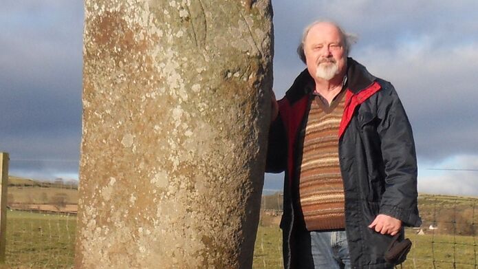 Alumnus Adrian C. Grant standing beside a tall ancient stone in a grassy field
