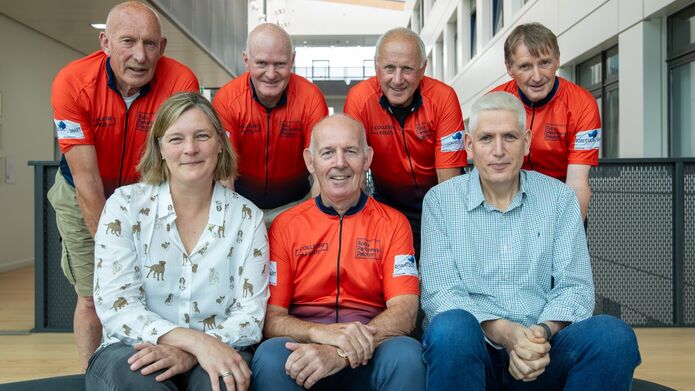 A group of seven people posing together indoors. Five are wearing orange cycling jerseys, while two sit at the front in casual clothing.
