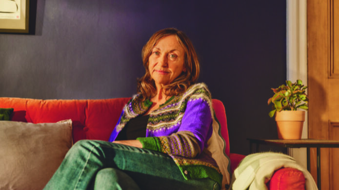 A woman sitting on a red sofa in a warmly lit room, looking towards the camera.