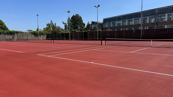An outdoor tennis court, with buildings and trees in the background and a bright blue sky.