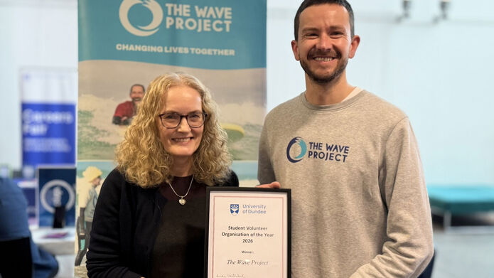 Two people holding a student volunteer prize certificate standing in front of a banner with the text The Wave Project 