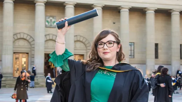 Photo of Aimee Hutchinson outsite Caird Hall wearing her graduation gown and waving her degree about