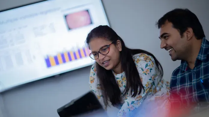 Students compare data on a laptop with a large screen behind them showing charts