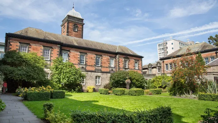 a red stone building with grass, seating and hedges out the front