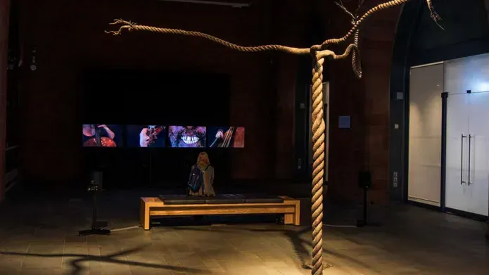 rope twisted to look like a tree, in a dark gallery with a screen showing people playing stringed instruments