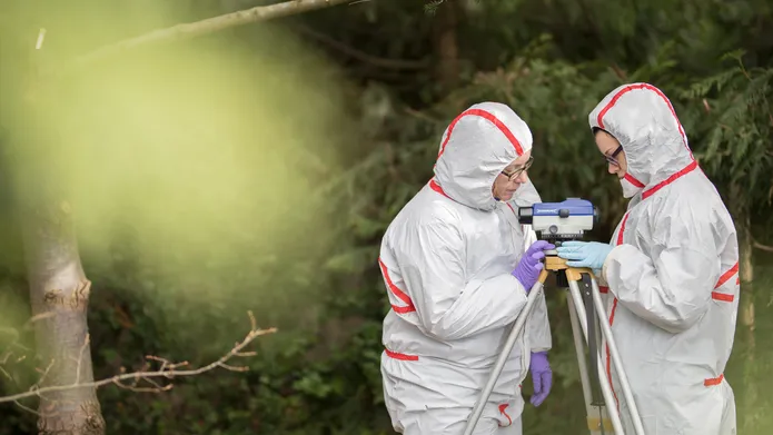 two people in full body clean suits look at a cameralike device on a tripod in the woods