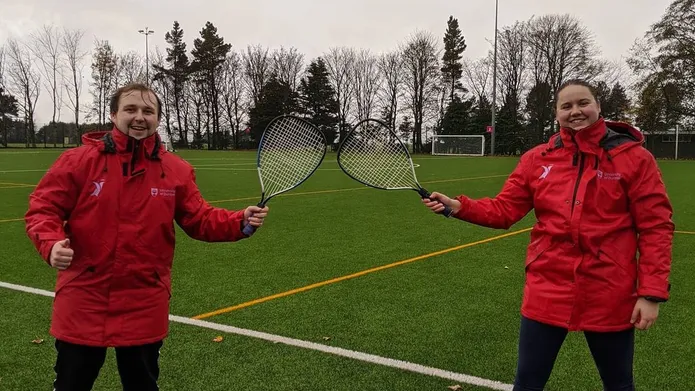 full shot of president dan and club member wearing red rain jackets standing on green grass pitch holding tennis rackets