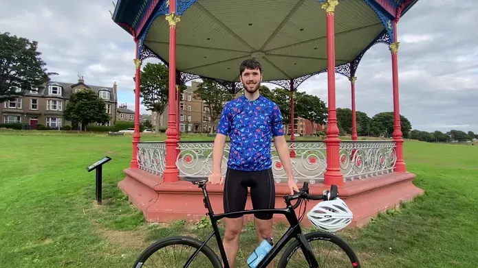 male in early 20's wearing tight blue and black cycling wear posing with black frame bike, standing in front of bandstand with red posts, surrounded by vibrant green grass