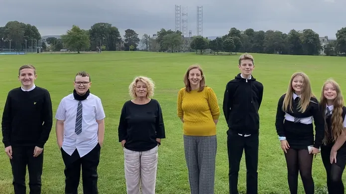 a group of five schoolchildren wearing uniform standing each side of two middle aged women in a park of vibrant green grass